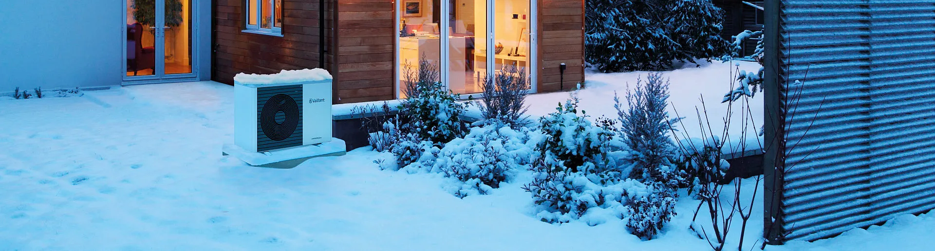Ein schneebedeckter Garten mit einer Vaillant-Wärmepumpe, die an der Außenwand eines Hauses installiert ist.