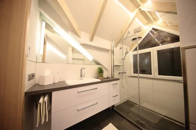 Modernes Dachbad mit sichtbaren Holzbalken, Waschtisch mit Spiegel und großer Glasdusche vor Dachfenster.