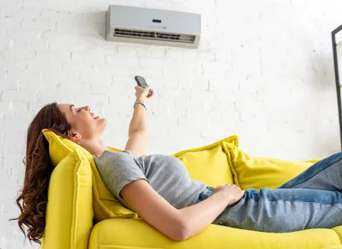 Eine Frau liegt entspannt auf einem leuchtend gelben Sofa und richtet eine Fernbedienung auf die Klimaanlage an der Wand. 