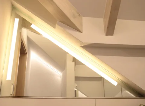 Detailaufnahme einer Dachschräge im Badezimmer mit sichtbaren Holzbalken und integrierter LED-Beleuchtung am Spiegel.