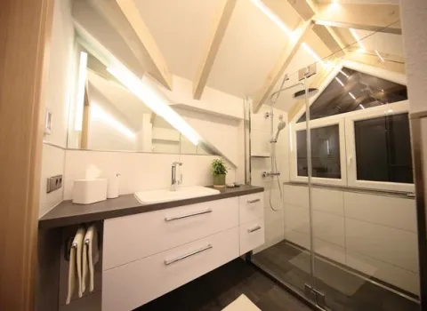 Modernes Dachbad mit sichtbaren Holzbalken, Waschtisch mit Spiegel und großer Glasdusche vor Dachfenster.