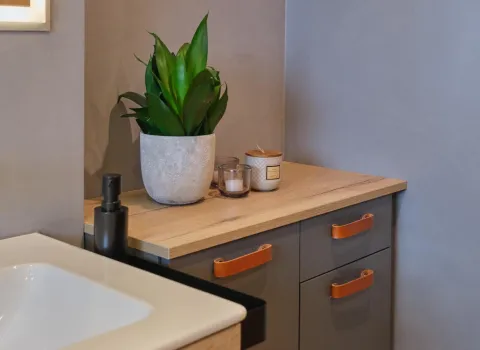 Detailansicht eines grauen Badmöbels mit Holzarbeitsplatte, Lederschlaufen-Griffen und Dekoration aus Pflanze, Kerze und Spender.