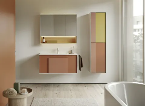 Ein Badezimmer im gerahmten Look zeigt Hängemöbel mit klar definierten weißen Rahmen, die farblich abgesetzte Fronten in Rostrot, Beige und Senfgelb einrahmen. Der Waschtisch und der hohe Seitenschrank wirken durch die Rahmenstruktur besonders strukturiert und hochwertig.
