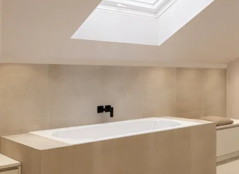 Eine moderne Badewanne ist unter einem großzügigen Dachfenster eingebaut und von beige-grauen Fliesen umgeben. Schwarze Wandarmaturen setzen einen eleganten Kontrast. Das Licht von oben sorgt für eine helle, entspannte Atmosphäre.
