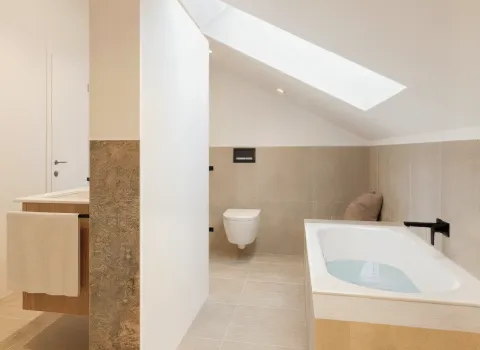 Ein helles Dachgeschossbad zeigt eine halb gefüllte Badewanne mit schwarzer Armatur und ein wandhängendes WC. Ein langes Dachfenster bringt natürliches Licht in den Raum. Dezente Beigetöne und klare Linien prägen das Design.