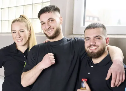 Drei junge Auszubildende Anlagenmechaniker posieren in Arbeitskleidung vor dem Werkstattsfenster.