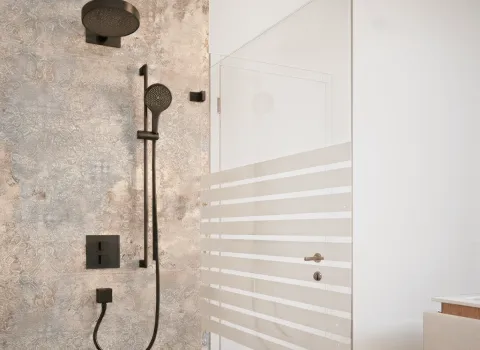 Ein modernes Duschkonzept mit schwarzer Regendusche, passender Handbrause und markanter Steinoptik-Wand. Der Duschbereich wird durch eine transparente Glaswand mit Streifendekor visuell abgetrennt. Die Ausstattung wirkt hochwertig und funktional.