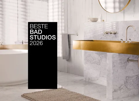 Ein stilvolles Bad mit goldenen Armaturen, Marmorwaschtisch und freistehender Badewanne. Ein schwarzer Banner mit "Beste Badstudios 2026" ist prominent im Bild.
