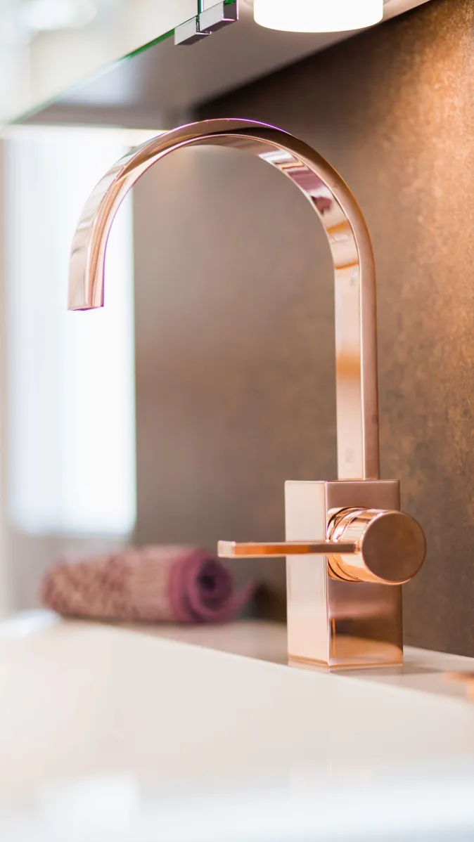 Detailaufnahme einer eleganten roségoldenen Badarmatur mit geschwungenem Auslauf, vor dunkler Wandfläche und dekorativem Handtuch.