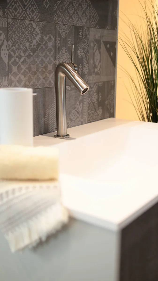 Modernes Waschbecken mit Edelstahlarmatur, grauer Fliesenrückwand im Musterdesign, dekoriert mit Handtuch, Seifenspender und Pflanze.
