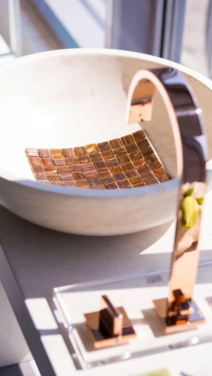 Rundes Steinwaschbecken mit Mosaikboden und eleganter roségoldener Armatur, ausgestellt in modernem Badambiente.