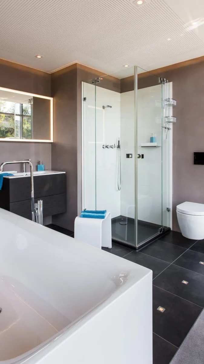 Modernes Badezimmer mit freistehender Badewanne, Glasdusche, wandhängendem WC, Waschtisch mit LED-Spiegel und dunklen Bodenfliesen.