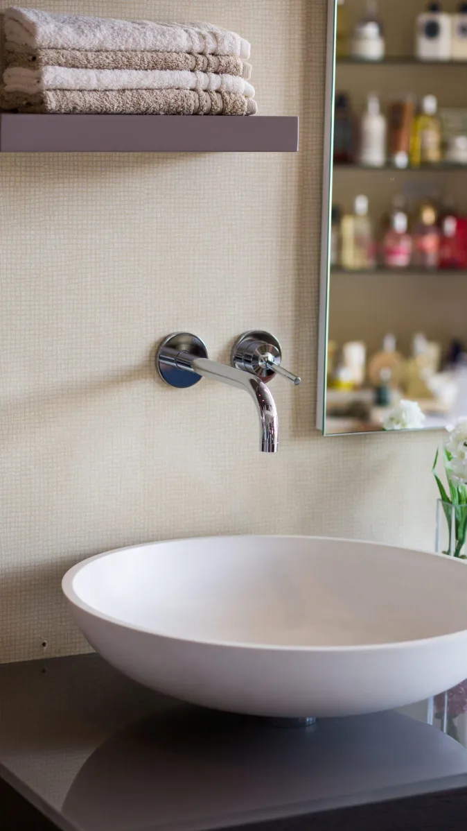 Rundes Aufsatzwaschbecken mit Wandarmatur, darüber gefaltete Handtücher auf Regal, daneben Spiegel und Blumendeko.