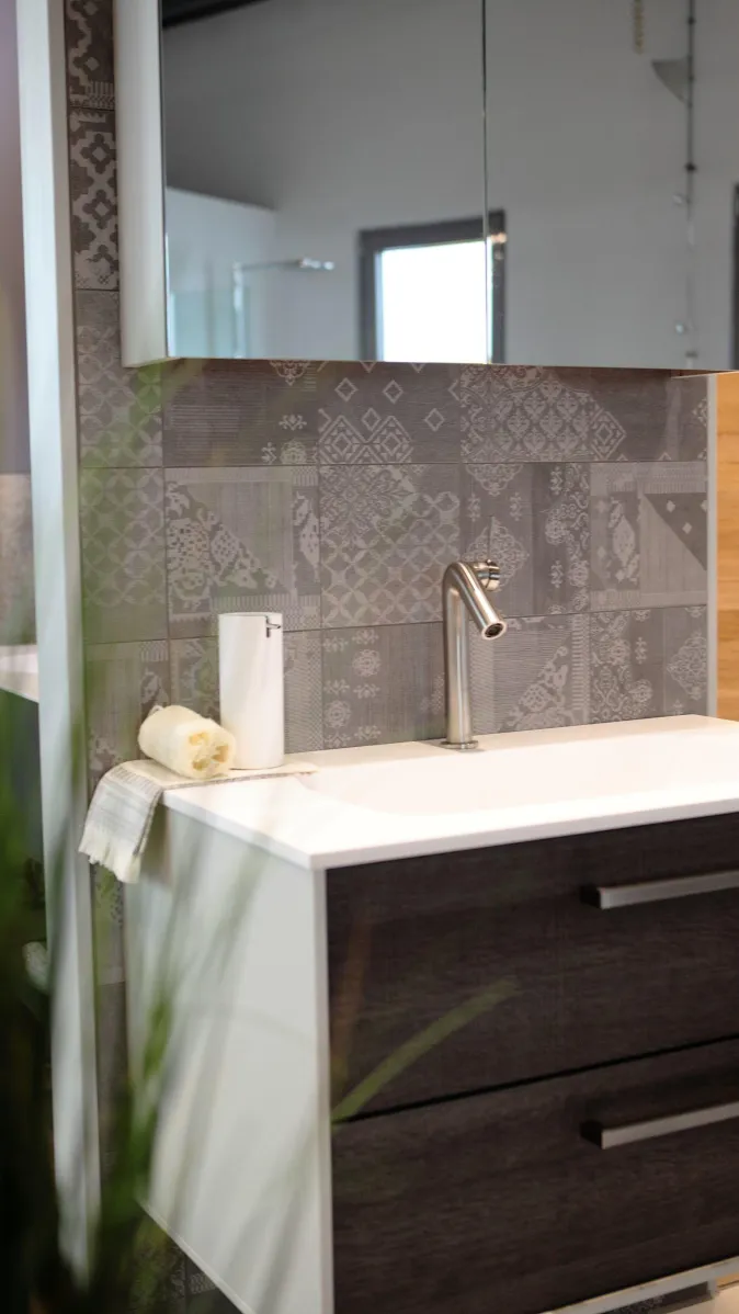 Moderner Waschtisch mit grauem Unterschrank, Edelstahlarmatur, Spiegel und Rückwand aus dekorativen Fliesenmustern.