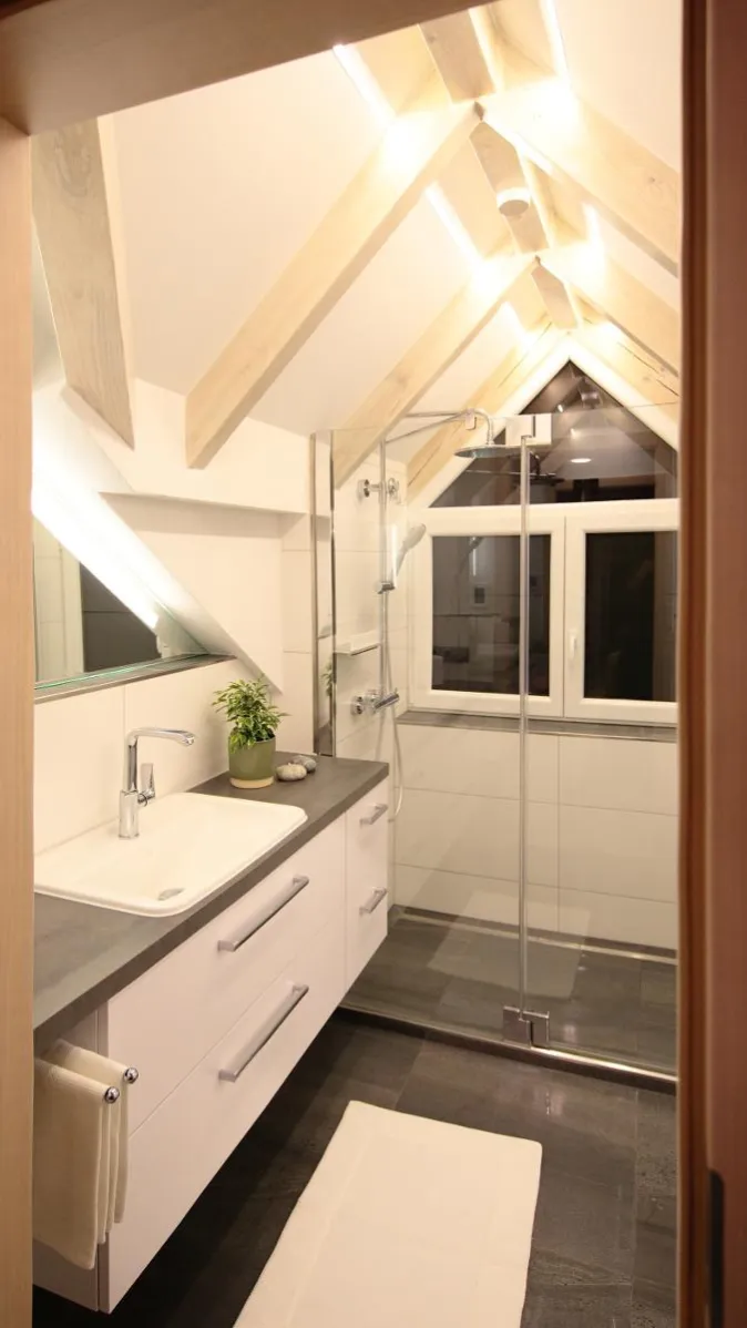 Modernes Dachbad mit Waschtisch, großem Spiegel, Glasdusche vor Fenster und sichtbaren Holzbalken mit LED-Beleuchtung.