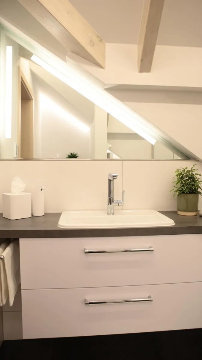 Waschtisch im Dachbad mit weißem Unterschrank, eingelassenem Waschbecken, moderner Armatur, Spiegel unter der Dachschräge und Pflanzendeko.