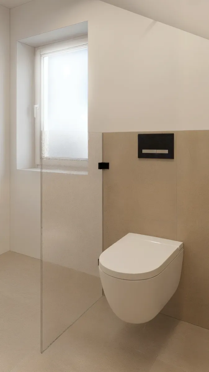 Ein modernes Badezimmer im Dachgeschoss mit einem wandhängenden WC vor beigen Fliesen. Eine matte Glasabtrennung trennt den WC-Bereich von einem Fenster mit Milchglas. Die schräg verlaufende Decke verleiht dem Raum architektonischen Charakter. Die minimalistische Gestaltung unterstreicht das moderne Design des Badezimmers.