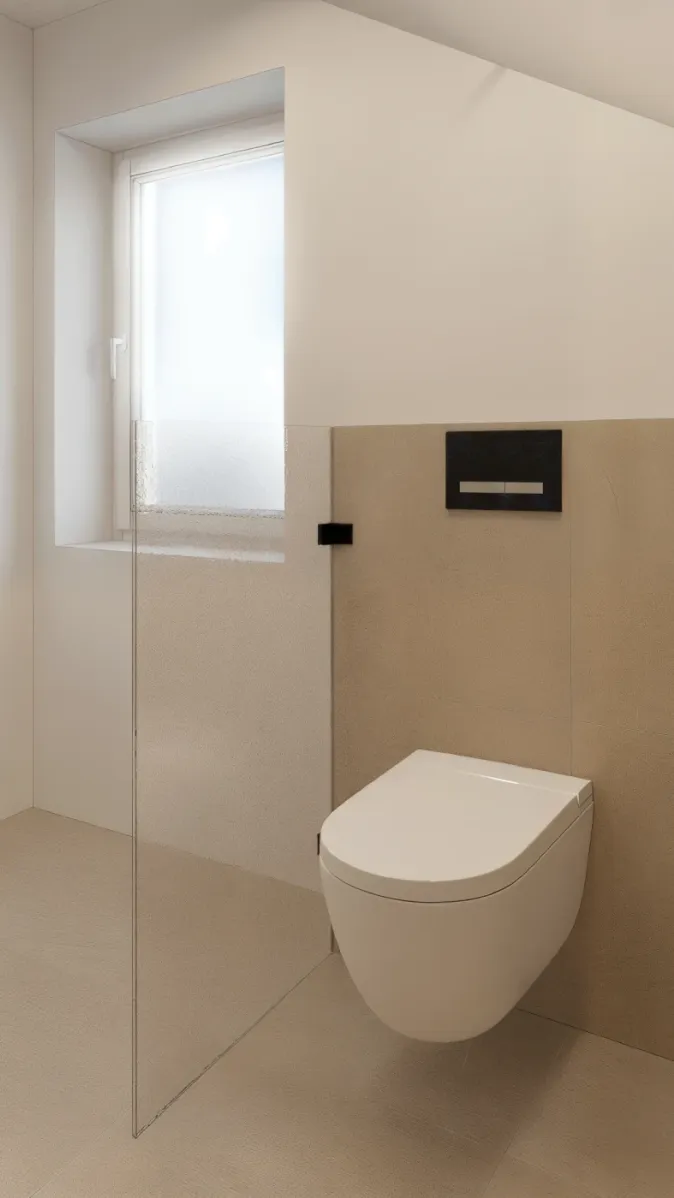 Ein modernes Badezimmer im Dachgeschoss mit einem wandhängenden WC vor beigen Fliesen. Eine matte Glasabtrennung trennt den WC-Bereich von einem Fenster mit Milchglas. Die schräg verlaufende Decke verleiht dem Raum architektonischen Charakter. Die minimalistische Gestaltung unterstreicht das moderne Design des Badezimmers.