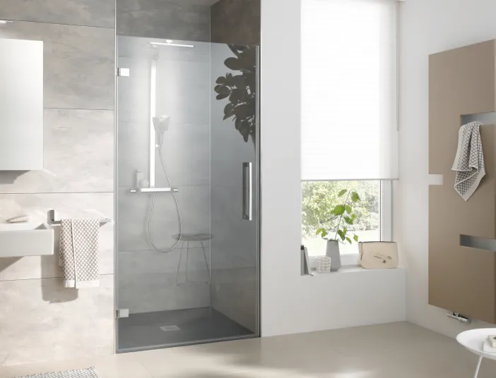 Helles Bad mit in Nische eingebauter Glasdusche HSK Atelier Plan, beige-grauen Fliesen, schlichtem Waschtisch und stilvollem Handtuchheizkörper vor einem großen Fenster.