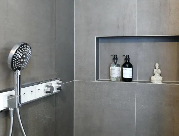 Moderne Dusche mit Handbrause, Thermostat-Bedienelement und Ablagefach mit Seifenspendern und kleiner Buddha-Figur.