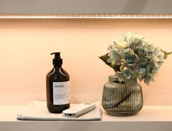 Detailaufnahme eines Wohnbads mit Seifenspender, Fernbedienung und Glasvase mit dekorativer Blume auf beleuchteter Ablage.