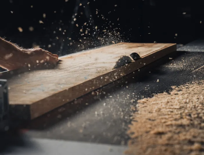 Ein Handwerker schneidet ein massives Holzbrett zu. Dabei fliegen Spaene durch die Luft.