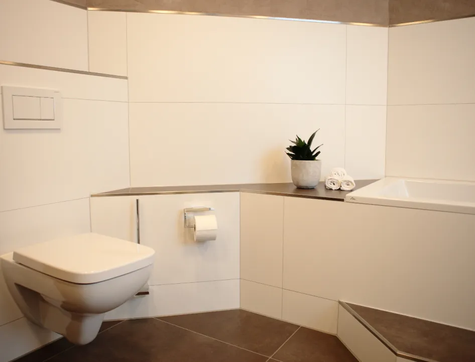 Modernes Bad mit Wand-WC, seitlicher Badewanne und dekorativer Ablage mit Pflanze und Handtüchern.