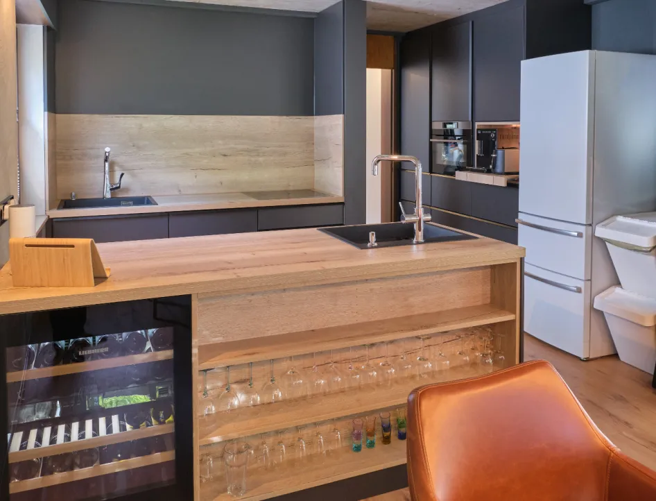 Moderne Wohnküche mit Kochinsel aus Holz, integriertem Weinkühlschrank, Gläserregal und schwarzen Küchenschränken.
